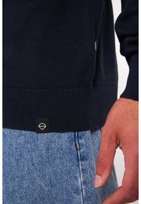 Dunkelblauer, gestrickter Pullover mit geripptem Saum, kombiniert mit hellblauen Jeans. Der Pullover hat ein kleines schwarzes Logo-Tag an der Seite.