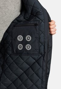 Hand die een zwarte doorgestikte jas opent en een binnenzak toont met iconen voor waterafstotend, winddicht, ademend en temperatuurbestendig.