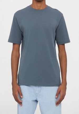 Mężczyzna ubrany w jednolitą, krótkim rękawem, ciemnoszarą koszulkę z okrągłym dekoltem i jasnoniebieskie dżinsy, stojący na jednolitym tle.