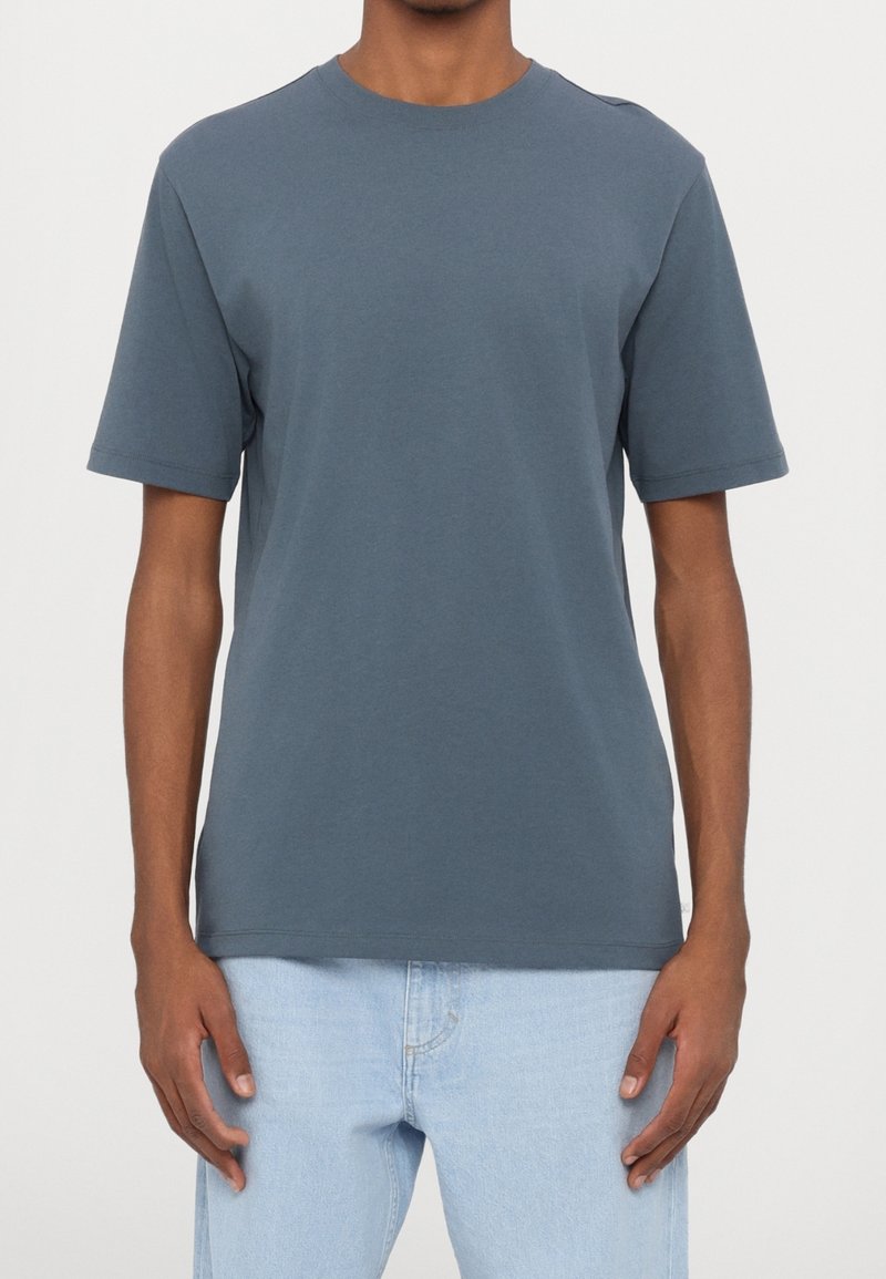 Homme portant un t-shirt gris foncé uni à manches courtes et col rond, et un jean bleu clair, debout devant un fond uni.