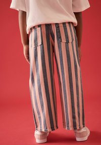 Jeans de pierna ancha a rayas en denim azul y rosa con bolsillos traseros. El modelo lleva zapatillas beige. El fondo es de color rojo sólido.