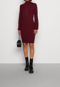 Robe en maille côtelée bordeaux avec une silhouette ajustée, un col montant et des manches longues. Associée à des bottines noires et un petit sac à main.
