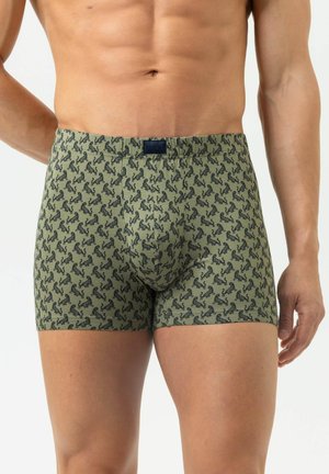 Mann trägt olivgrüne Boxershorts mit einem kleinen schwarzen Tiermotiv vor weißem Hintergrund.