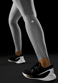 Szare legginsy z teksturowymi panelami i bocznymi akcentami. Czarne buty sportowe z białym obcasem i detalem marki, umieszczone na ciemnym tle.