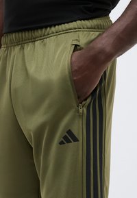 Σκοτεινός άνθρωπος που φοράει παντελόνι φόρμας Adidas σε ελιά πράσινο με μαύρες ρίγες και λογότυπο, με το χέρι μέσα στην τσεπή με φερμουάρ στο πλάι.