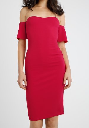 Mujer con un vestido rojo ajustado, sin hombros, hasta la rodilla y con mangas cortas que caen por debajo de los hombros, sobre un fondo liso.