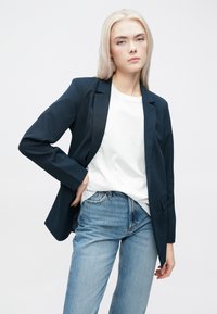 Jūrų laivyno švarkas su pritaikytu dizainu, su įpjovimais ant atviro krūtinės, ir vieno mygtuko fasadų uždarymu, derinamas su balta marškinėliais ir šviesiai mėlynais džinsais.