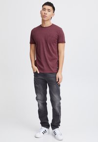 Bordeauxrotes T-Shirt mit kurzen Ärmeln und Brusttasche, kombiniert mit ausgewaschenen, schwarzen Jeans und weißen Sneakers mit schwarzen Streifen. Schlichte, lässige Passform.