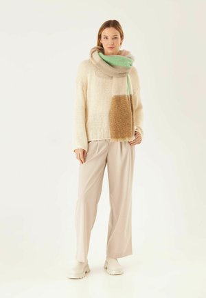 Crèmekleurige gebreide trui en losse beige broek; een multicolor sjaal in zachtgroen, bruin en crèmekleurig om de nek gedrapeerd. Witte chunky schoenen.