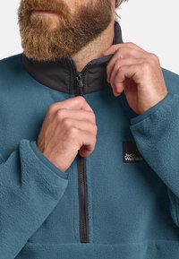 Halve-rits fleece pullover in teal met een zwarte kraag. Kenmerken zijn een gestructureerde afwerking en een logo patch op de linkerbovenkant van de borst, versterkt ritsgebied.