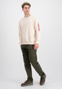 Sudadera de cuello redondo beige con detalle de cremallera lateral y etiqueta roja; combinada con pantalones cargo verde oliva y zapatillas marrones.