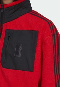 Chaqueta de forro polar roja con un bolsillo negro con cremallera y un cuello negro en contraste. Presenta tres rayas negras en el hombro.