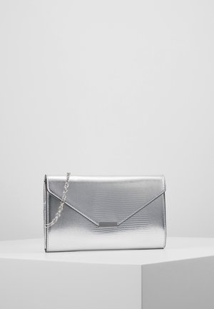 Sølv metalisk tekstureret clutch taske med konvolutlåg og tynd kæderem, der hviler på en hvid piedestal mod en lysegrå baggrund.