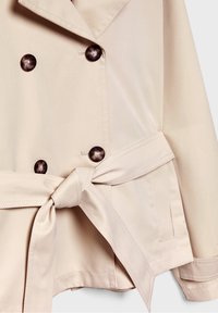 Beige trench coat with double-breasted design, large dark buttons, tied belt, and visible stitching details on the collar and cuffs.