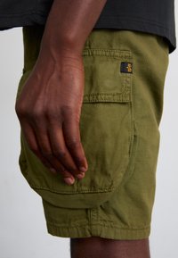 Olive green cargo shorts with a textured fabric, featuring a side pocket and a small black tag with an orange logo. Hand resting on the shorts.