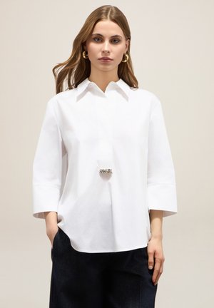 Biała bluzka z kołnierzykiem i rękawami o długości trzech ćwierć, ozdobiona kryształowym detalem przy dekolcie, wykonana z gładkiej bawełnianej tkaniny.
