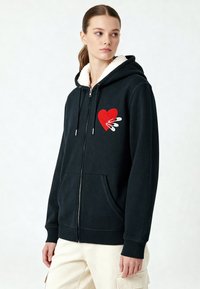 Zwarte rits hoodie met een fleece-gevoerde kraag. Kenmerkt zich door een rood hartontwerp met witte accenten op de borst en de voorkeurszakken. Soepele textuur.