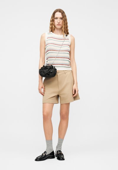 Γυναίκα με κυματιστά μαλλιά φοράει αμάνικο ριγέ τοπ, μπεζ σόρτς, γκρι κάλτσες, μαύρα loafers και κρατά μαύρη τσάντα με λουλούδια και αλυσίδα.