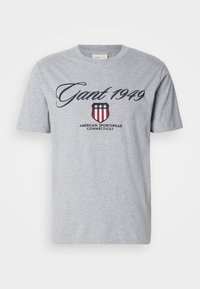 Camiseta de algodão cinza com mangas curtas, apresentando "Gant 1949" em letra cursiva e um logotipo com um escudo, estrelas e riscas.