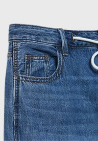 Gros plan sur un jean en denim bleu montrant la poche avant, la poche à monnaie, la passants de ceinture et le cordon blanc à la taille sur un fond clair.
