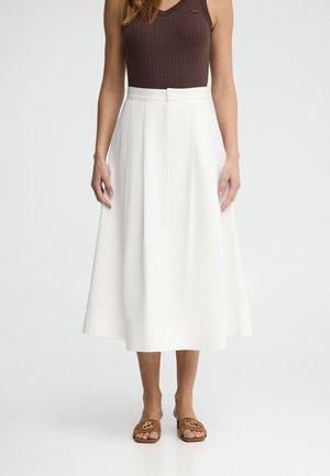 Kvinde iført brun ærmeløs ribbet top, hvid A-formet midinederdel med høj talje og brune åbne sandaletter, står på hvid baggrund.