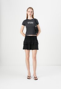 Black cropped t-shirt with white text, paired with a black tiered mini skirt. Accessories include a small handbag and black flip-flops.