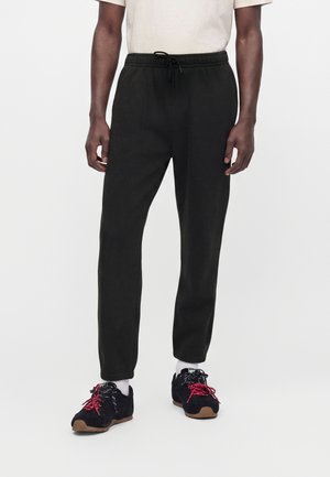 Mann trägt schwarze Kordelhose, weißes Hemd, weiße Socken und schwarze Sneakers mit roten Schnürsenkeln und steht vor einem schlichten weißen Hintergrund.