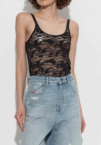 Donna che indossa un top nero in pizzo abbinato a jeans di denim chiaro a vita alta e strappati, in piedi contro uno sfondo semplice.