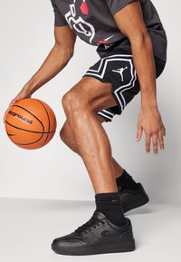 Pantalones cortos deportivos negros con líneas de acento blancas, zapatillas negras, camiseta gris y un balón de baloncesto naranja sostenido en una pose dinámica.