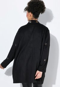 Chaqueta de estilo ante negra con un corte relajado, que presenta ojales de metal en las mangas, un respaldo recto y un brillo sutil.