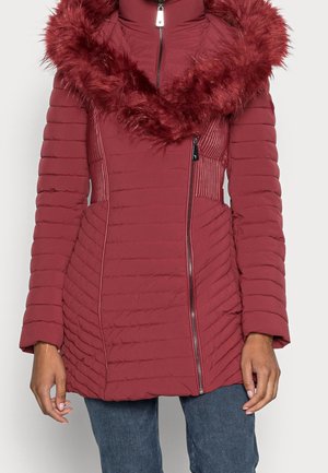 Vrouw draagt een gewatteerde, bordeauxrode jas met een diagonale ritssluiting en een grote namaakbontkraag, gecombineerd met een blauwe spijkerbroek.