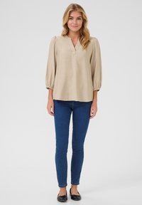 Blouse beige, ample avec des manches ballons 3/4 et un décolleté en V, associée à un jean bleu slim et des ballerines noires. Texture lisse.