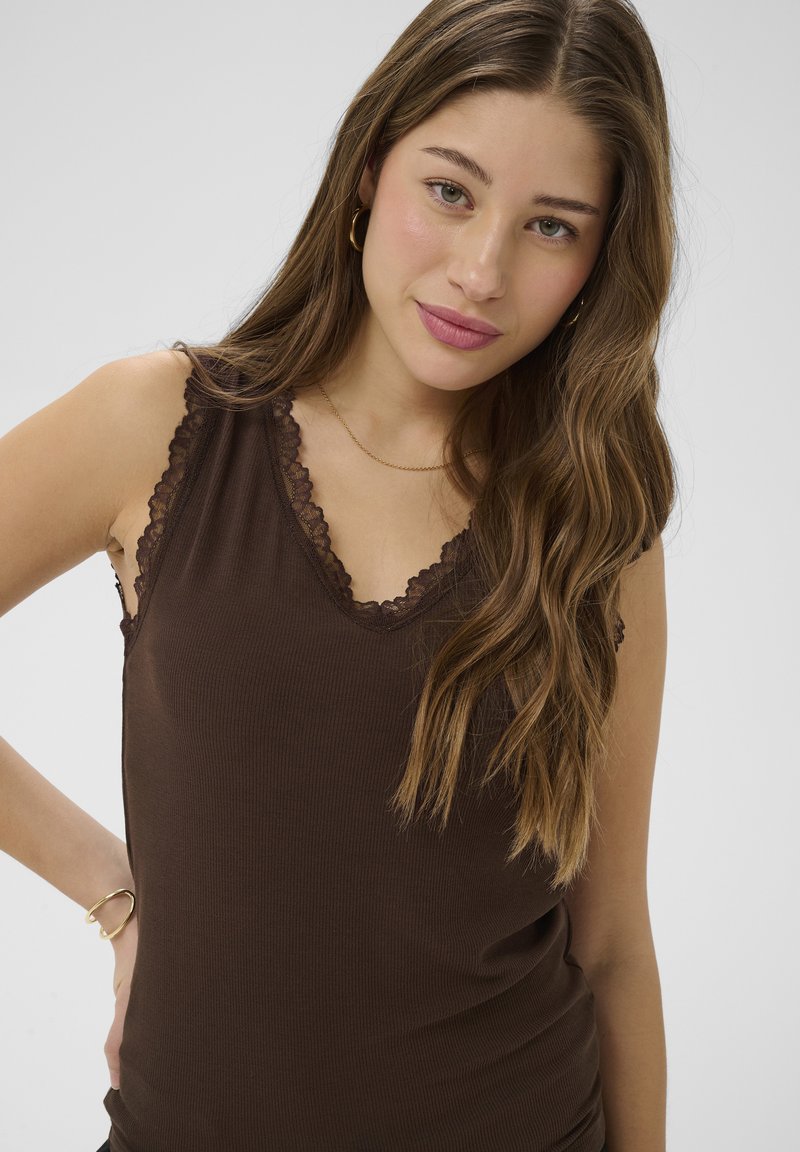 Jeune femme aux longs cheveux bruns ondulés portant un haut sans manches marron foncé orné de dentelle, des boucles d'oreilles anneaux dorés et un bracelet.