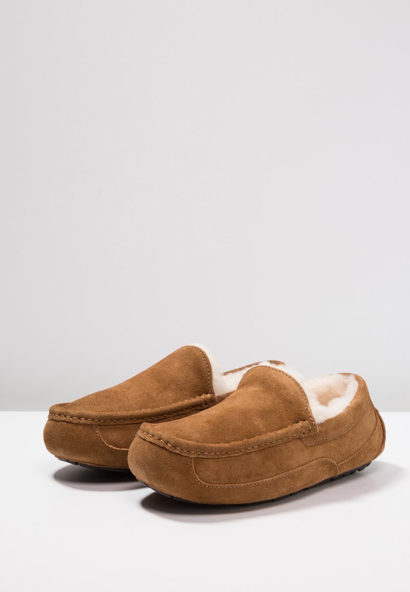 Brown suede slippers with a soft, white fleece lining, featuring a slip-on design and visible stitching detail. Flat rubber soles.