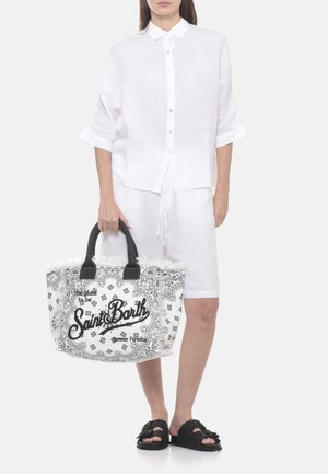 Donna che indossa una camicia bianca con bottoni e pantaloncini, che tiene una grande borsa bianca con motivo paisley nero e manici neri.