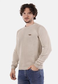 Maglione lavorato a maglia beige con scollatura rotonda, maniche lunghe e un sottile dettaglio del logo sul petto, abbinato a jeans blu.