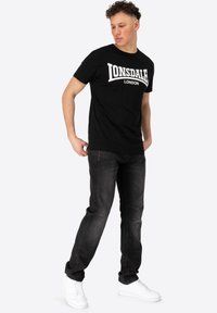 Juodas medvilnės marškinėliai su balta „Lonsdale London“ logotipu, derinami su tamsiomis džinsinėmis kelnių ir baltais sportiniais bateliais. Modelis stovi šiek tiek pasilenkęs.