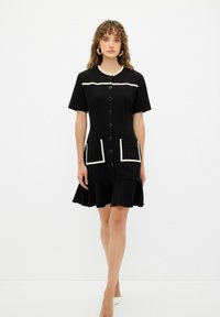 Vestido negro con mangas cortas, dos bolsillos frontales y ribete blanco. Forma en A con un dobladillo con volantes y cierre de botones en la parte delantera.