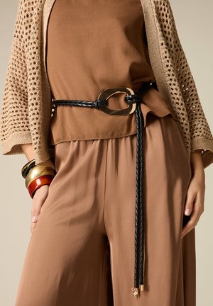 Femme portant un pantalon ample marron et un top avec un cardigan beige en maille, accessoirisée d'une ceinture noire tressée et de plusieurs bracelets épais.