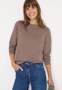 Maglia a maniche lunghe marrone in tessuto morbido, vestibilità rilassata, scollatura rotonda, indossata con jeans blu, mano in tasca, che mette in risalto uno stile casual.