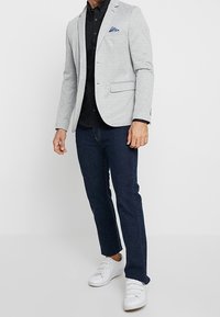 Hombre con blazer gris claro y pañuelo azul en el bolsillo, camisa negra, vaqueros azul oscuro y zapatillas blancas, posando frente a un fondo blanco.