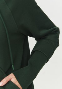 Sudadera verde oscuro con un corte relajado, que cuenta con capucha con cordón, puños acanalados y un bolsillo tipo canguro. Textura de tela suave.