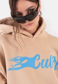 Beżowa bluza z kapturem z grafika w niebieskie płomienie. Model ma także na sobie czarne okulary przeciwsłoneczne. Gładka tekstura, luźny krój i minimalne detale szwów.