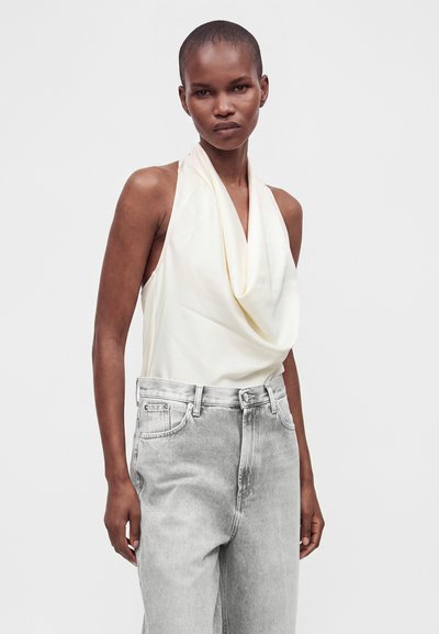 Femme aux cheveux courts portant un top blanc sans manches à col bénitier et un jean gris clair taille haute sur un fond blanc uni.