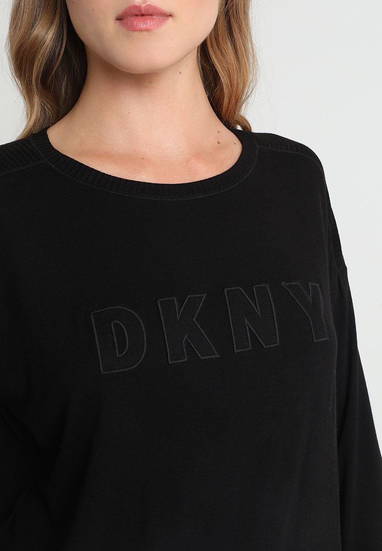 Kobieta w czarnym swetrze z wypukłymi, teksturowanymi literami "DKNY" na piersiach oraz okrągłym dekoltem.