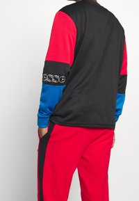 Man iklädd en svart, röd och blå färgblockerad sweatshirt med matchande röda byxor, med händerna i fickorna, vänd bort från kameran.