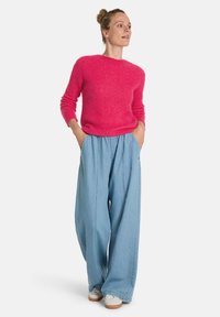 Rosa gestrickter Pullover mit langen Ärmeln, kombiniert mit hellblauen, weit geschnittenen Jeans. Das Model steht mit den Händen in den Taschen vor einem weißen Hintergrund.