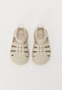 Beige sandals with a closed toe and open sides, featuring horizontal straps, textured soles, and a rear heel loop for easy wear.