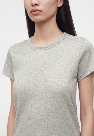 Femme aux longs cheveux foncés portant un t-shirt gris clair uni à manches courtes et col rond sur fond blanc.
