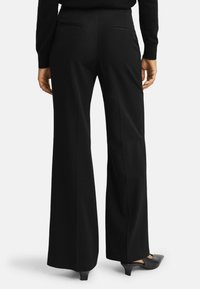 Zwarte, wijde broek met een gladde stofafwerking, voorzien van twee achterzakken en een getailleerde pasvorm, gecombineerd met zwarte schoenen met een lage hak.